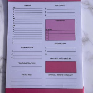 May include: A pink and white daily planner with gold spiral binding. The planner includes sections for scheduling, high-priority tasks, goals, actions, and affirmations. The words "DAILY PLANNER" are printed at the top.