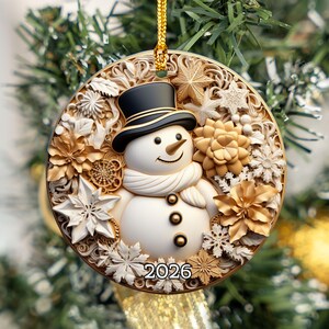 May include: A round Christmas ornament featuring a central white snowman wearing a black top hat and scarf, surrounded by intricate gold and white floral and snowflake patterns, with the year 2026 printed in black at the bottom center.