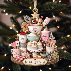 Könnte beinhalten: Ein Weihnachtsbaumschmuck mit Back-Motiv, bestehend aus Miniatur-Küchenutensilien, Cupcakes und Gefäßen mit den Aufschriften "Baked with Love", "Made with Love", "Sugar", "Flour" und "Happiness is Homemade". Der Sockel ist mit dem Namen "SOPHIA" personalisiert.