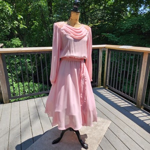 May include: A pink vintage dress with a cowl neck and long sleeves. The dress has a sash at the waist and is made of a sheer fabric. The dress is on a mannequin and is displayed against a wooden background.