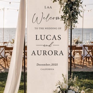 Puede incluir: Un letrero de bienvenida de boda con los nombres Lucas y Aurora, y la fecha de diciembre de 2025, en California. El letrero está decorado con flores blancas y vegetación, con una tela drapeada a un lado.