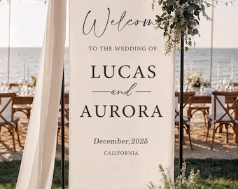 May include: A wedding welcome sign with the names Lucas and Aurora, and the date December 2025, in California. The sign is decorated with white flowers and greenery, with a draped fabric on the side.