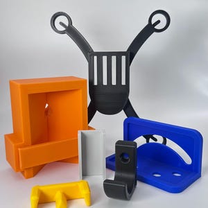 May include: An assortment of 3D-printed objects in various colors, including orange, yellow, black, white, and blue. The objects include a box, a hook, a bracket, and a support structure. The items are arranged against a white background.