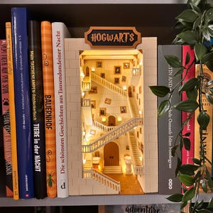 Könnte beinhalten: Ein Miniatur-Bücherregal-Einsatz mit einem detaillierten Innenraum einer Schlosstreppe mit mehreren Ebenen, weißen Geländern und kleinen gerahmten Porträts an den Wänden. Die Struktur ist von innen beleuchtet und trägt oben das Schild HOGWARTS.