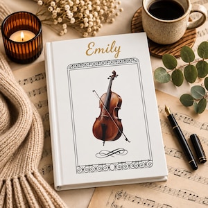 May include: A white hardback book featuring the name Emily in gold script at the top. The cover displays a detailed illustration of a brown cello with a bow inside a black rectangular frame with decorative scrollwork borders at the top and bottom.