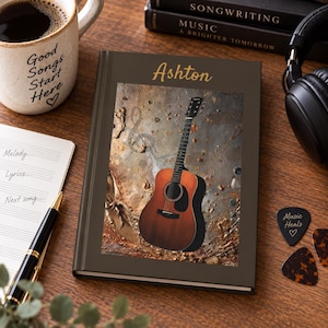 May include: A dark brown hardcover journal featuring the name Ashton in gold script at the top and a central rectangular image of an acoustic guitar against a textured metallic background. The journal is positioned on a wooden surface.