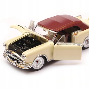 Op de afbeelding: Een crèmekleurig vintage cabriomodel met een bordeauxrood dak en open deuren. De motorkap is open en onthult de motor. De auto heeft chromen accenten en whitewall-banden. Een reservewiel is zichtbaar aan de achterkant.