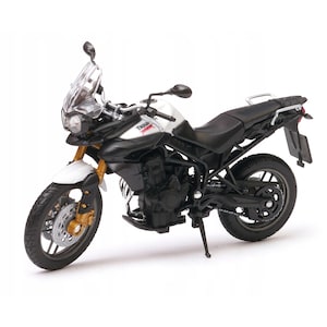 Può includere: Modello dettagliato di una motocicletta Triumph Tiger 800. La moto presenta una combinazione di colori bianco e nero con dettagli dorati sulle forcelle anteriori e sulle ruote. Il modello include parabrezza, specchietti e cavalletto.