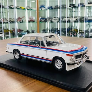 May include: A white scale model car featuring a two-door coupe body style with a distinctive blue and red racing stripe running along the side and across the hood. The vehicle includes silver wheels, chrome bumpers, and a black interior visible through windows.