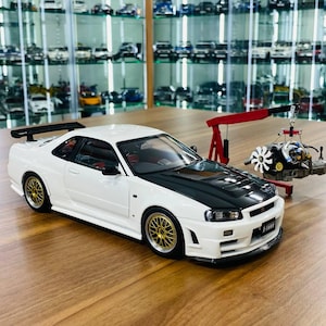 May include: A white and black scale model sports car with gold multi-spoke wheels and a rear spoiler. Next to the car is a red miniature engine hoist lifting a detailed car engine with a white cooling fan, all displayed on a wooden surface.