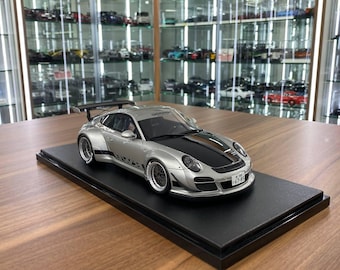 May include: A silver and black scale model sports car featuring a rear wing, wide fenders, and a Japanese license plate reading 67-85. The vehicle is mounted on a rectangular black display base, showcasing detailed wheels and a sleek aerodynamic body.