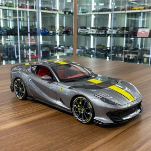 Könnte beinhalten: Ein graues Sportwagen-Modellauto mit einem gelben Rennstreifen auf der Motorhaube und dem Dach. Das Fahrzeug hat eine rote Innenausstattung, schwarze Räder mit gelben Felgenakzenten und ein gelbes Ferrari-Emblem an der Seitenverkleidung.