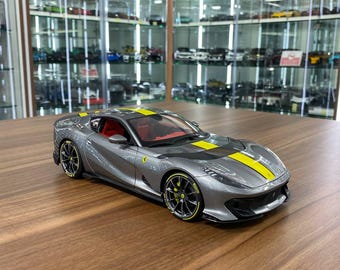 May include: A gray scale model sports car featuring a yellow racing stripe running along the hood and roof. The vehicle has red interior seating, black wheels with yellow rim accents, and a yellow Ferrari emblem on the side panel near the door.