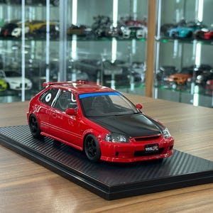 Op de afbeelding: Een rood model van een Honda Civic Type R hatchback met een zwarte motorkap, zwarte wielen en een blauwe zonneband op de voorruit. Het voertuig staat op een zwarte basis met koolstofvezeltextuur en een Civic Type R kentekenplaat.