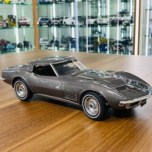 May include: A metallic dark grey scale model of a Chevrolet Corvette Stingray sports car. The miniature vehicle features a sleek aerodynamic body, chrome-trimmed wheels with white-walled tyres, a black interior, and clear windows showing the cabin.