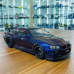 以下が含まれることがあります： リアスポイラー、ブラックのマルチスポークホイール、GT-Rエンブレム付きの精巧なフロントグリルを備えたダークブルーのNissan Skyline GT-R R34スケールモデルカー。光沢のある仕上げ、クリアなヘッドライト、空力特性に優れたボディが特徴です。