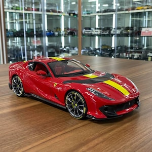 May include: A red scale model sports car featuring a yellow racing stripe running along the hood and roof. The vehicle has black multi-spoke wheels with yellow rim accents, a low-profile aerodynamic body, and a visible red interior cabin.