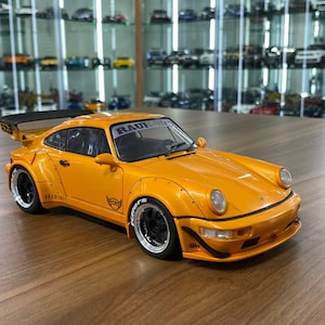 May include: A bright orange scale model sports car featuring a wide body kit, a large black rear spoiler, and black wheels with white lettering on the tires. The windshield displays the text RAUH-Welt, and the side panels feature black decals.