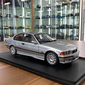 May include: A silver scale model of a two-door coupe car featuring a detailed exterior with multi-spoke wheels, orange turn signal indicators, and a front license plate. The miniature vehicle is mounted on a black rectangular display base.