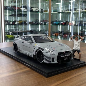 Może przedstawiać: Szary model samochodu sportowego Nissan GT-R z czarnymi kołami, dużym tylnym spojlerem i napisem LB Works na przednim grillu. Obok na czarnej podstawie stoi mała figurka osoby ubranej w biały t-shirt i ciemne szorty.