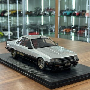 May include: A silver and black scale model car featuring a two-tone paint scheme, gold mesh wheels, and a black front grille. The vehicle displays the text RS-TURBO on the side and front license plate, and it is mounted on a black textured display base.