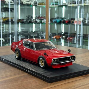 May include: A red scale model of a vintage coupe car featuring gold-rimmed wheels, black fender flares, and side-mounted mirrors. The vehicle is displayed on a rectangular black textured base, showcasing a detailed front grille and headlights.