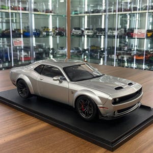 May include: A silver scale model of a Dodge Challenger muscle car featuring wide fender flares, black wheels with red brake calipers, and a black interior. The vehicle is mounted on a rectangular black display base with a smooth finish.