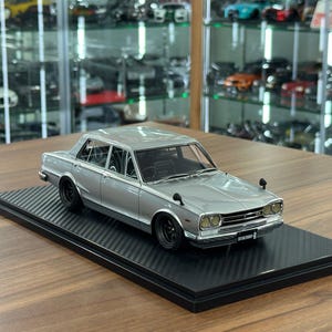 May include: A silver scale model of a Nissan Skyline GT-R saloon featuring black wheels, wing-mounted side mirrors, and a front grille emblem. The miniature vehicle is displayed on a black rectangular base with a textured carbon fibre pattern.