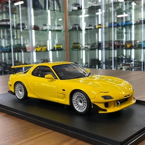 Puede incluir: Un modelo a escala de un coche Mazda RX-7 amarillo con alerón trasero, llantas plateadas de radios múltiples e interior negro. El vehículo en miniatura está montado sobre una base rectangular negra, mostrando líneas de carrocería detalladas.