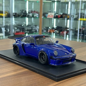 May include: A blue scale model sports car featuring a wide body kit, a large rear wing, and black wheels. The vehicle is mounted on a rectangular black display base, showcasing a detailed exterior with round headlights and a low-profile stance.