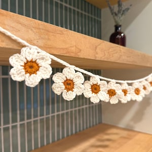 Könnte beinhalten: Eine gehäkelte Gänseblümchen-Girlande mit weißen Blütenblättern und orangefarbenen Zentren. Die Girlande ist auf einer weißen Schnur aufgereiht und wird auf einem Holzregal präsentiert. Der Hintergrund zeigt eine grüne geflieste Wand.