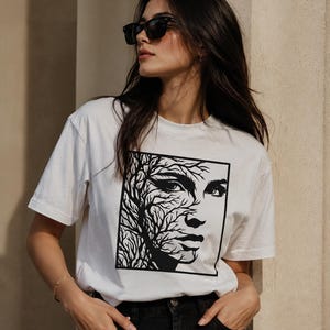 Peut inclure: Un t-shirt blanc à manches courtes avec une impression graphique carrée noire sur la poitrine. Le motif représente un visage humain partiellement fusionné avec des branches d'arbre complexes. Le design est centré sur le devant du vêtement en coton.