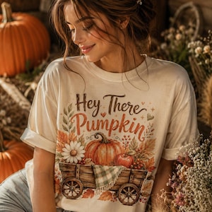 May include: A cream-coloured short-sleeve t-shirt featuring a graphic print of a wooden wagon filled with a pumpkin, an apple, and flowers. The text "Hey There Pumpkin" is printed above the wagon in a brown script font with small heart accents.