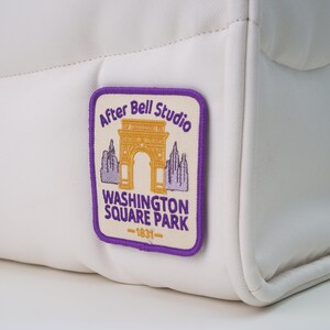 Puede incluir: Bolso blanco acolchado con un parche rectangular. El parche presenta el texto "After Bell Studio" sobre una ilustración del arco de Washington Square Park, con el texto "WASHINGTON SQUARE PARK -1931-" debajo.