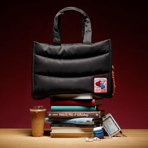 Puede incluir: Bolso tote negro acolchado con un parche que dice "One Big Kiss, I am Not a Thief" sobre una pila de libros. Una bebida de café, un teléfono y otros accesorios están junto a los libros. El fondo es rojo oscuro.