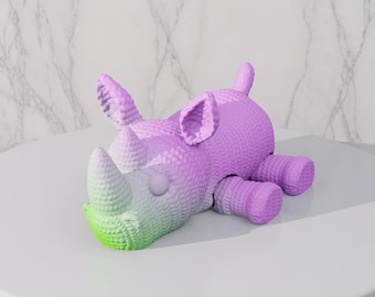 May include: A whimsical, knitted rhino toy in shades of purple and green. The rhino is lying down, with its horn and snout in a gradient of green, transitioning to purple. The body is a solid purple color, with detailed knitted texture.