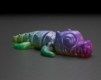 May include: A colorful, articulated alligator toy in shades of green, blue, purple, and red. The toy has a segmented body, with visible joints, and a large, expressive eye. The alligator is lying on a dark gray surface.