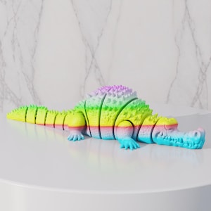 Peut inclure: Figurine d'alligator segmentée aux couleurs de l'arc-en-ciel. Le jouet alligator est posé sur une surface blanche. Les couleurs comprennent le jaune, le vert, le bleu et le violet. L'alligator a une colonne vertébrale texturée et est un objet décoratif unique.