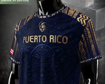 Puede incluir: Una camiseta azul marino con detalles dorados y el texto "PUERTO RICO". La camiseta presenta un diseño detallado con un escudo y un patrón texturizado. La tela es ligera y transpirable.