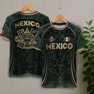 Op de afbeelding: Twee groene t-shirts met een gouden Azteekse kalenderontwerp en het woord "MEXICO" in gouden letters. Eén shirt toont de Mexicaanse vlag en de andere de vlag op de mouw. De shirts hangen aan houten hangers.