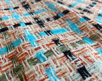 Op de afbeelding: Close-up van een geweven stof met een textuurpatroon. De stof heeft een mix van kleuren, waaronder oranje, blauw, zwart, groen en crème. Het ontwerp is een herhalend rasterachtig patroon.