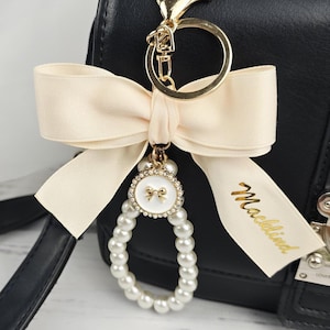 May include: A cream-colored bow keychain with a gold clasp and ring. The keychain features a pearl-beaded charm with a gold bow design and the word "Macklind" printed on a ribbon.
