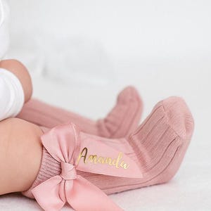 May include: A baby wearing a white long-sleeved onesie and pink knee-high socks. The socks have a bow detail and the name "Amanda" in gold script. The socks are ribbed and made of a soft material.