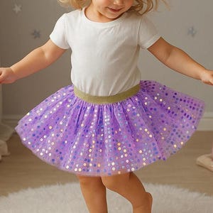 May include: A young child wearing a white t-shirt and a purple tutu skirt with gold sequins. The skirt has a gold waistband. The child is smiling and appears to be dancing. The background is a light gray wall with star-shaped decorations.
