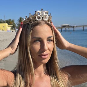 May include: A gold-colored crown with the number 35 in rhinestones. The crown is worn on a person's head. The person is wearing a white dress and a gold necklace. The background is a beach with a pier and blue water.