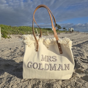 Ivory personalized bridal tote bag with “Mrs Goldman” pearl letters, fringe top, and leather handles photographed on a sandy beach under sunlight.