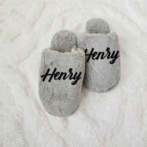 Close-up of fluffy grey slippers with the name “Henry” printed on top, placed on soft white blanket — custom men's gift.