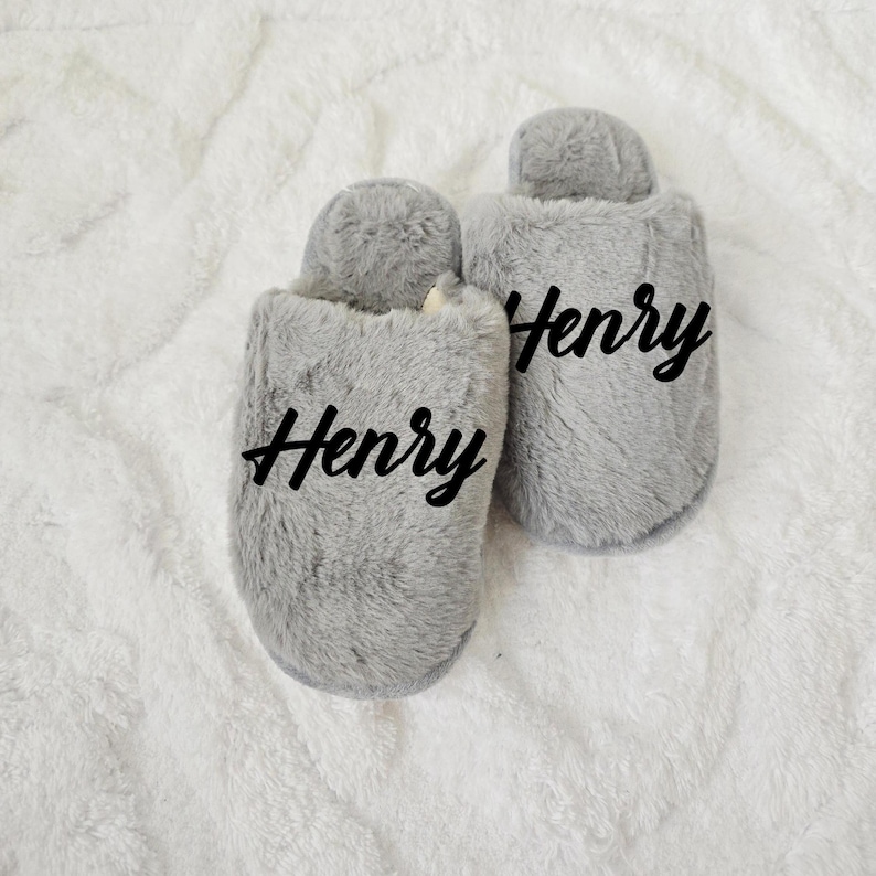 Close-up of fluffy grey slippers with the name “Henry” printed on top, placed on soft white blanket — custom men's gift.