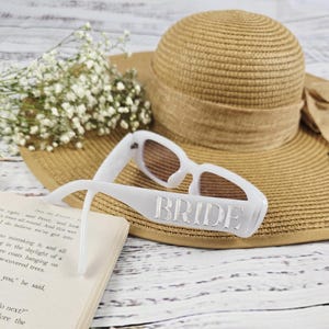 White BRIDE sunglasses beside a brown straw hat and book, surrounded by small flowers, perfect for bridal shower or honeymoon outfit inspiration.