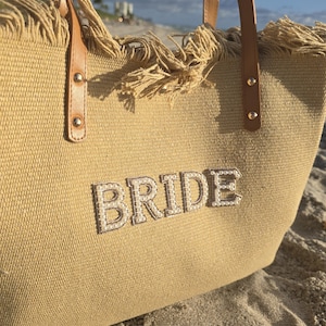 Close-up of beige bridal tote bag with pearl “BRIDE” letters and fringe detail, resting on sandy beach.